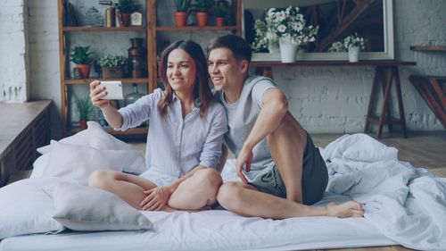Free A happy couple taking a selfie while sitting on a cozy bed in a stylish, plant-filled room. Stock Photo