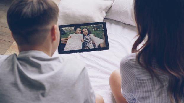 A couple watching a tablet screen showing a friends' photo while sitting on a bed.