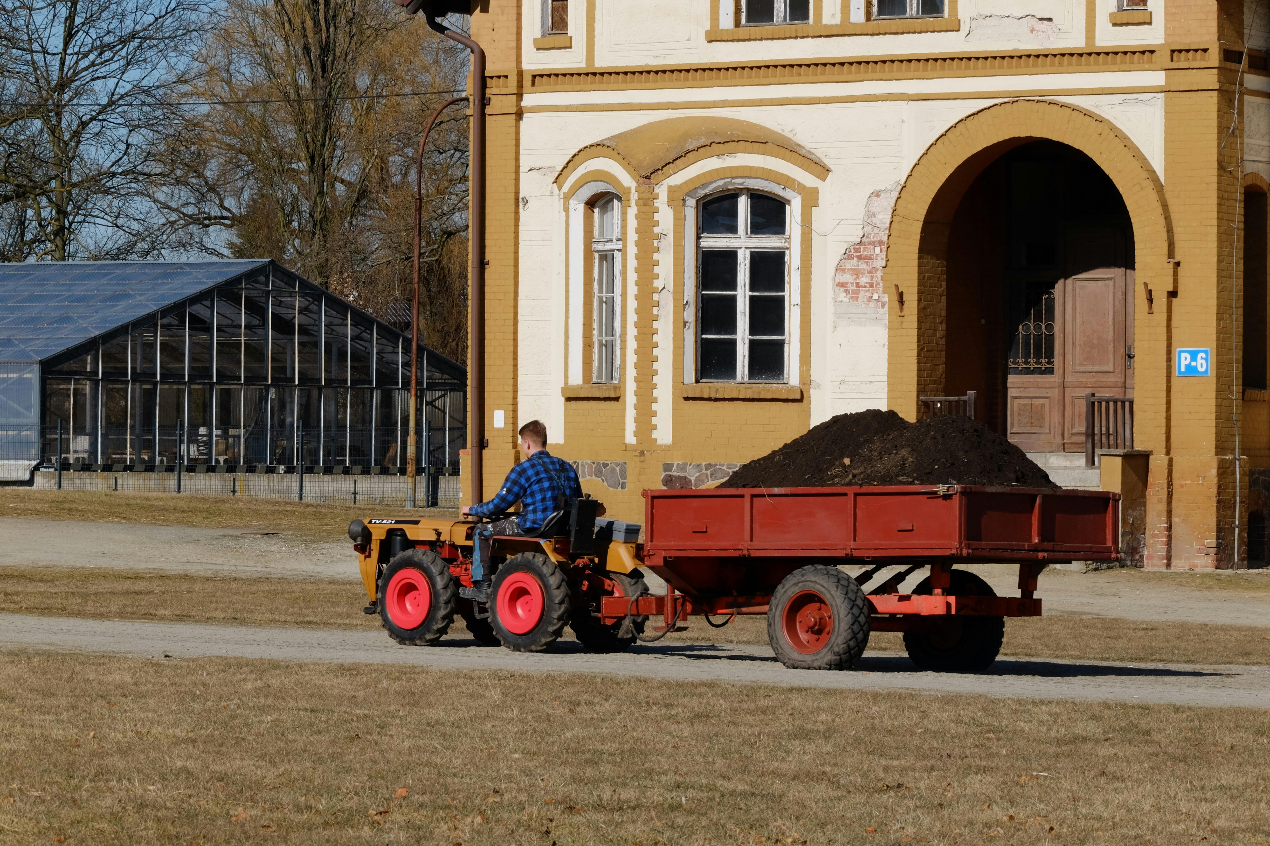 A gardener drives a tractor towing soil past a greenhouse and historic building.