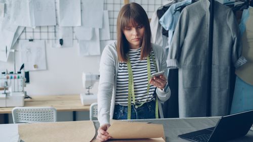 Free Young fashion designer checks smartphone while reviewing design patterns in her creative studio. Stock Photo