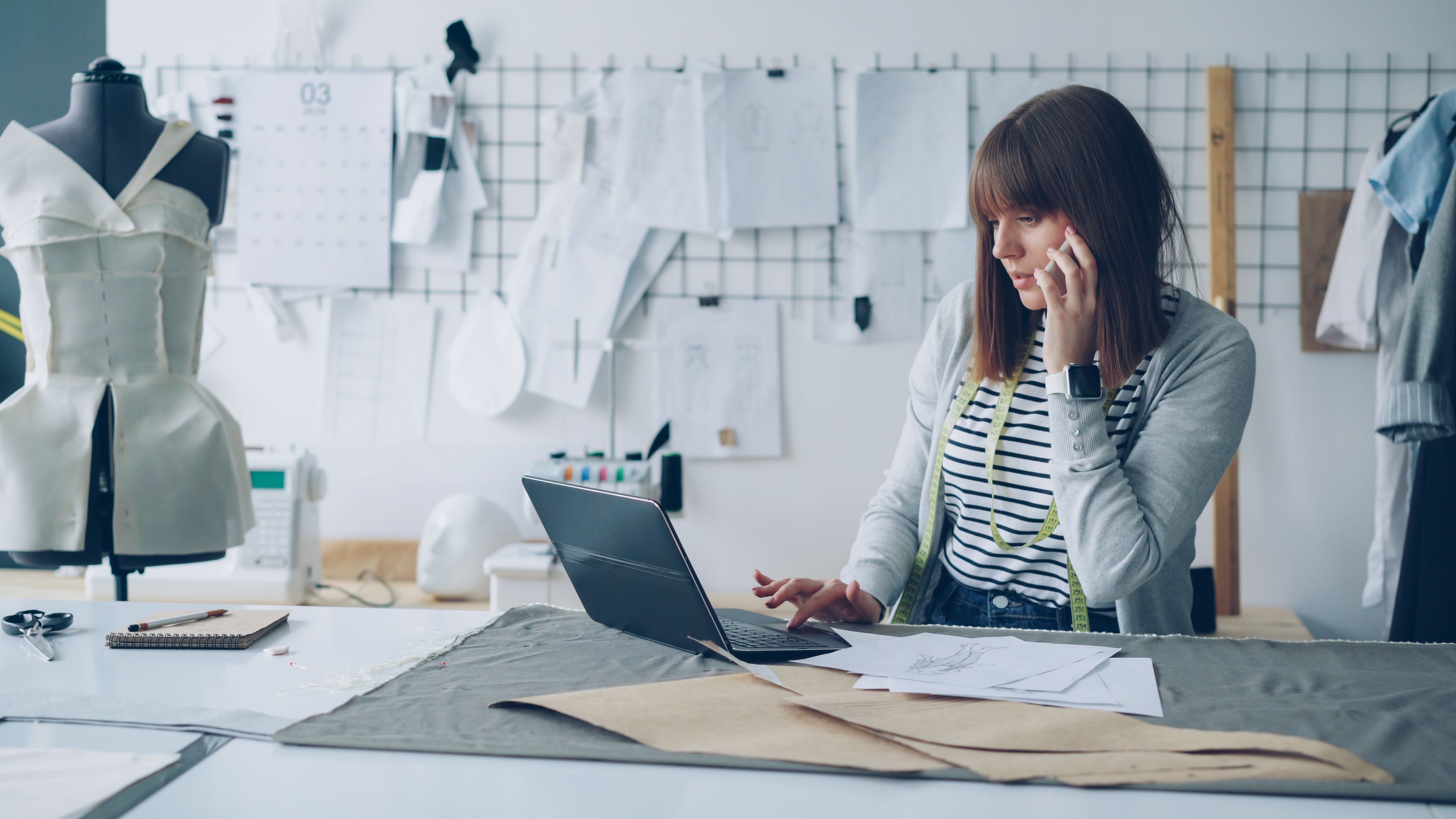 A female fashion designer working on her laptop in a creative design studio, surrounded by design sketches.