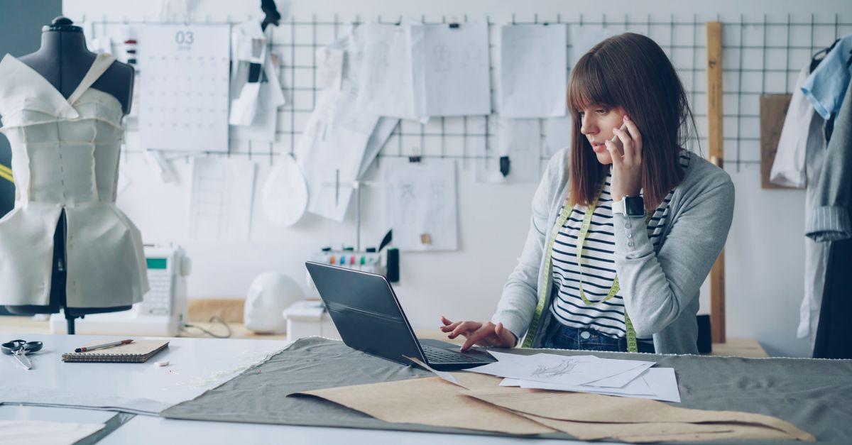 A female fashion designer working on her laptop in a creative design studio, surrounded by design sketches.