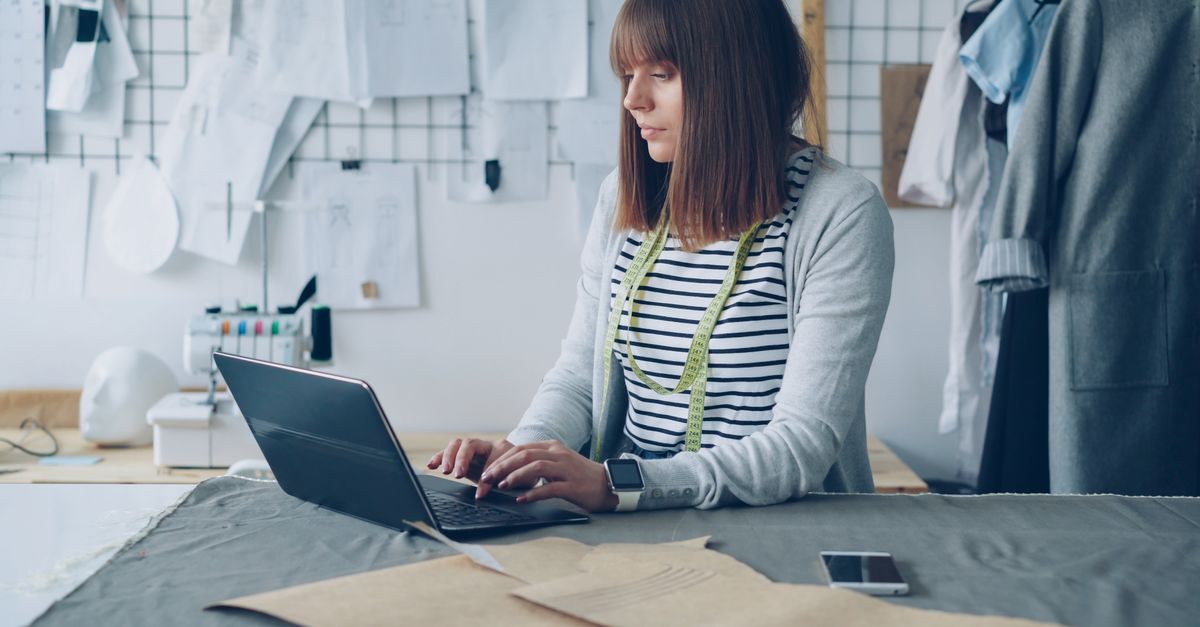Female fashion designer working on laptop in creative studio space, surrounded by sketches.