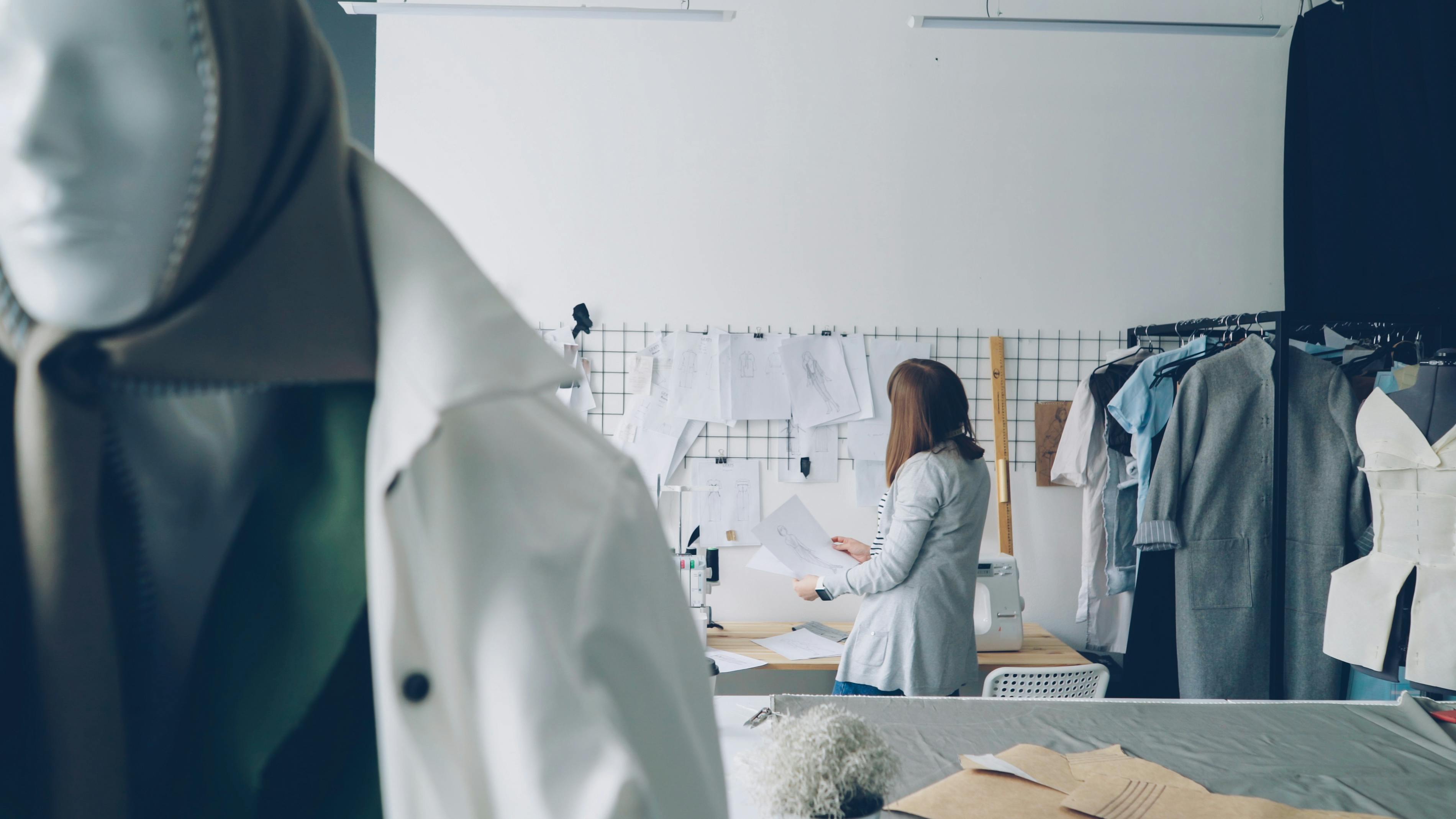 A fashion designer reviews sketches in a modern workspace with mannequins and patterns.