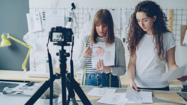 Two female fashion designers discussing and sharing creative clothing sketches in a studio setting.