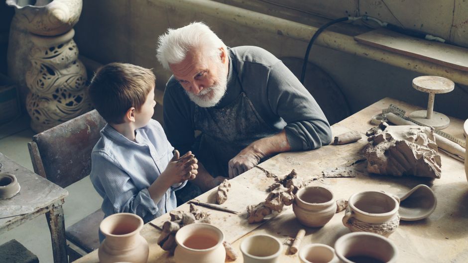 A senior man teaching pottery skills to a young boy in an artsy workshop setting.