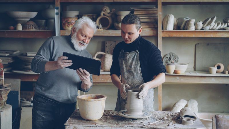 An instructor with a tablet observing a learner working at a potter's wheel