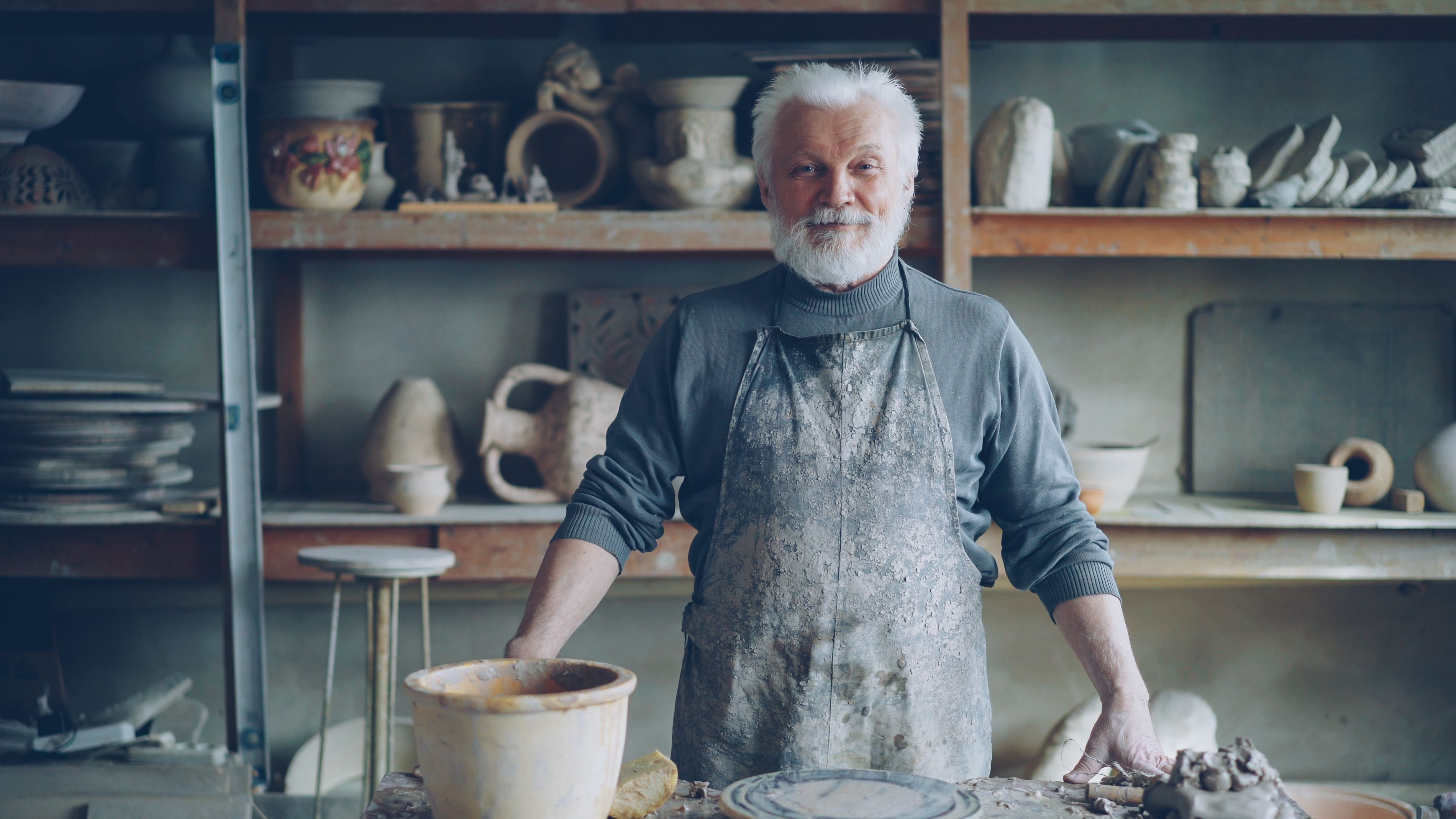 Elderly male potter in rustic ceramics studio surrounded by pottery.