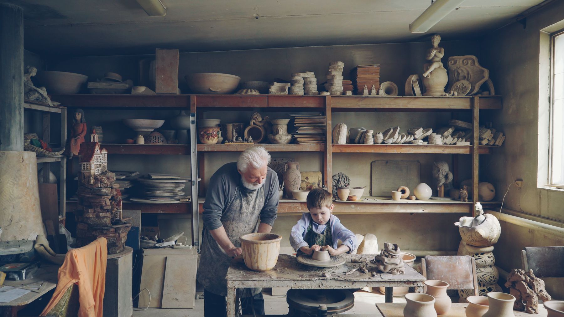 An older craftsman guiding a young apprentice at the wheel in a cluttered workshop
