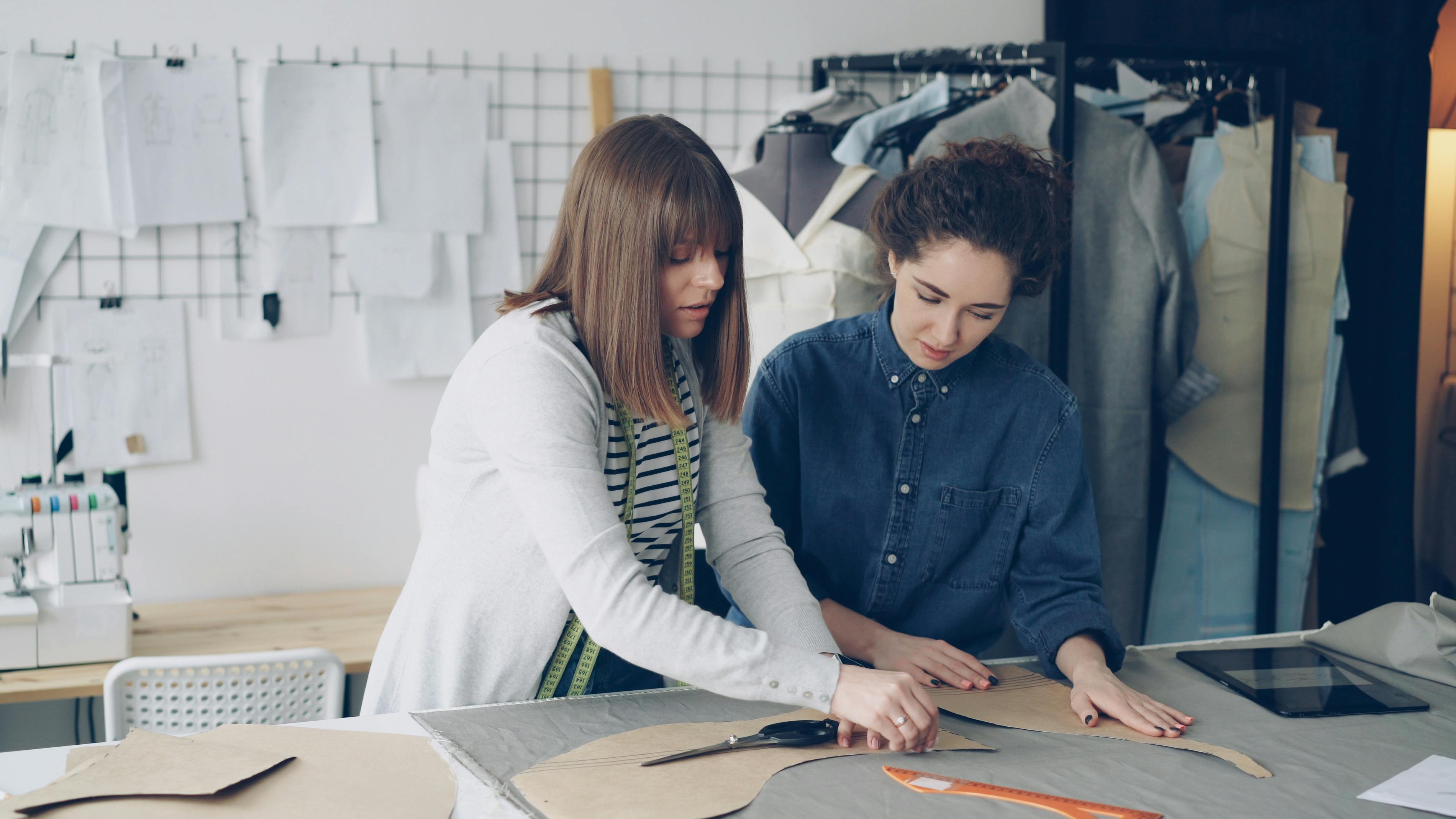 Two women work on fashion designs in a creative workshop environment.