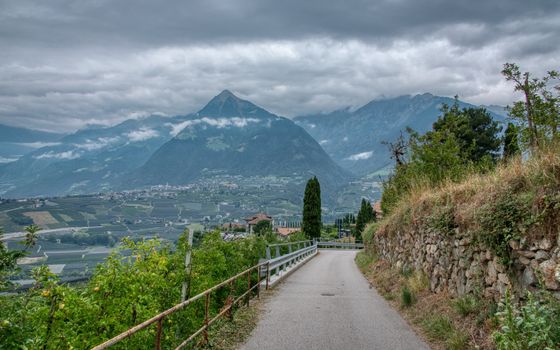 A picturesque road through Schenna, Italy, with stunning mountain and vineyard views under cloudy skies.