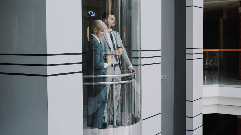 Two business professionals in suits standing inside a modern glass elevator.