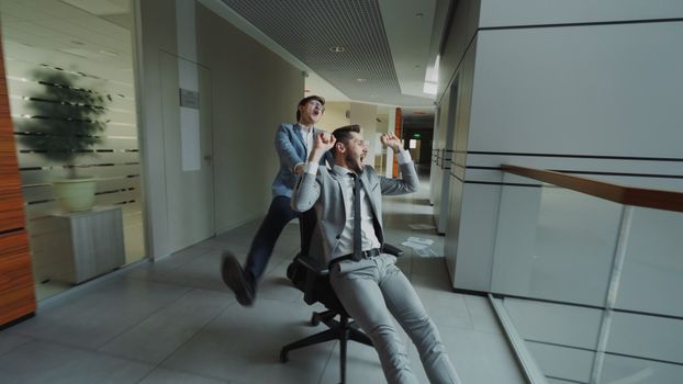 Two businessmen having fun racing in office chairs down a hallway.