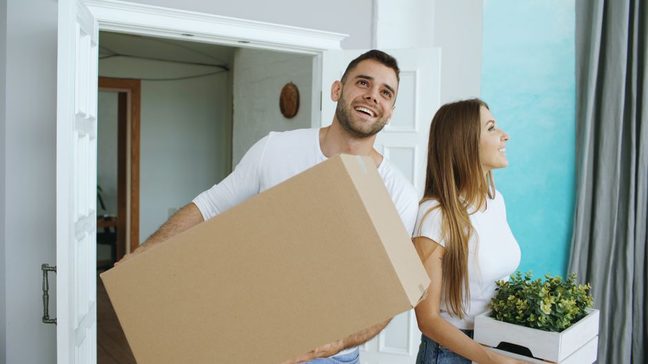 Una pareja alegre se muda a su nuevo hogar, cargando cajas y una planta.