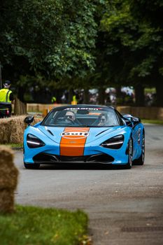 A vivid McLaren sports car races on a tree-lined track in Beaulieu, UK.