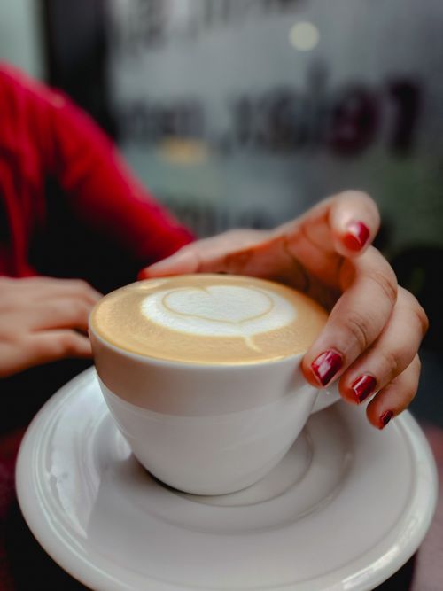 Free Close-up of hands with red nails holding a latte with heart-shaped art in a cafe. Stock Photo