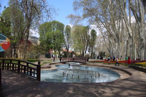 Free A beautiful park scene featuring a wooden bridge over a fountain surrounded by trees and flowers. Stock Photo
