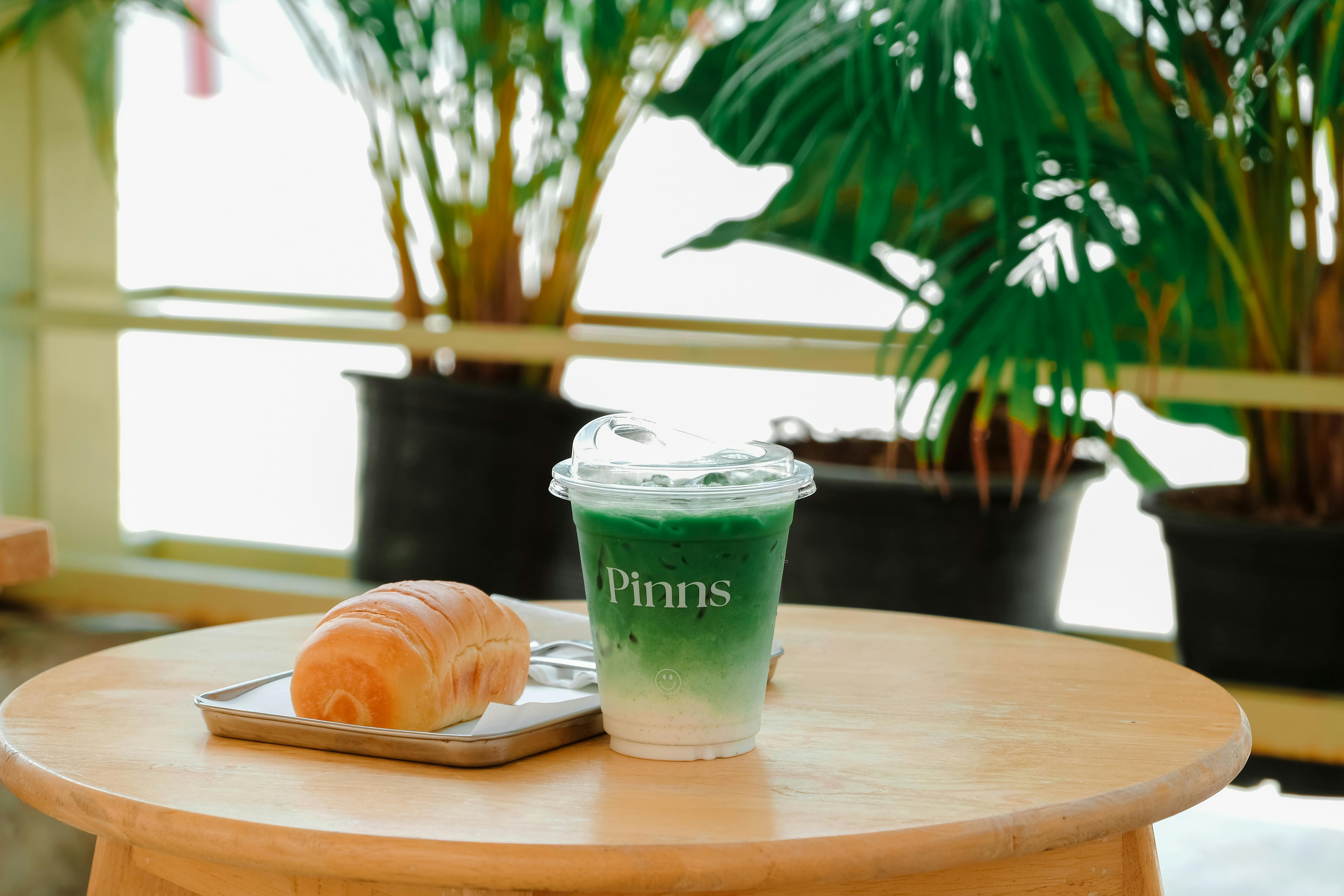 Green iced matcha latte and fresh croissant on a wooden table in a cozy cafe setting with indoor plants.