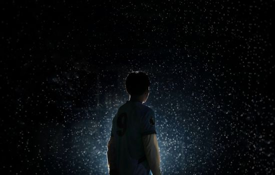 A dramatic silhouette of a soccer player standing in rain with backlit lighting.
