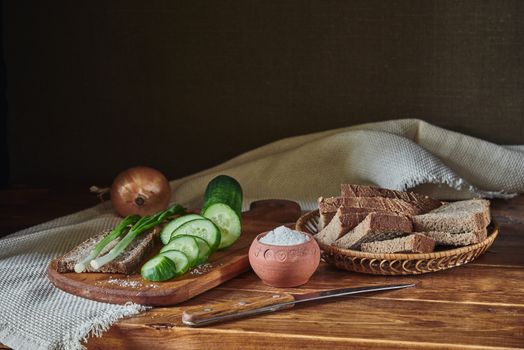 A rustic arrangement of bread, cucumbers, onion, and salt on a wooden table.