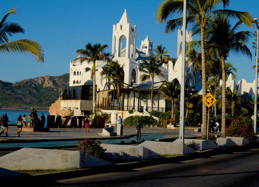 Scenic view of Mazatlán's iconic seaside with people, architecture, and palm trees under the sunny sky.