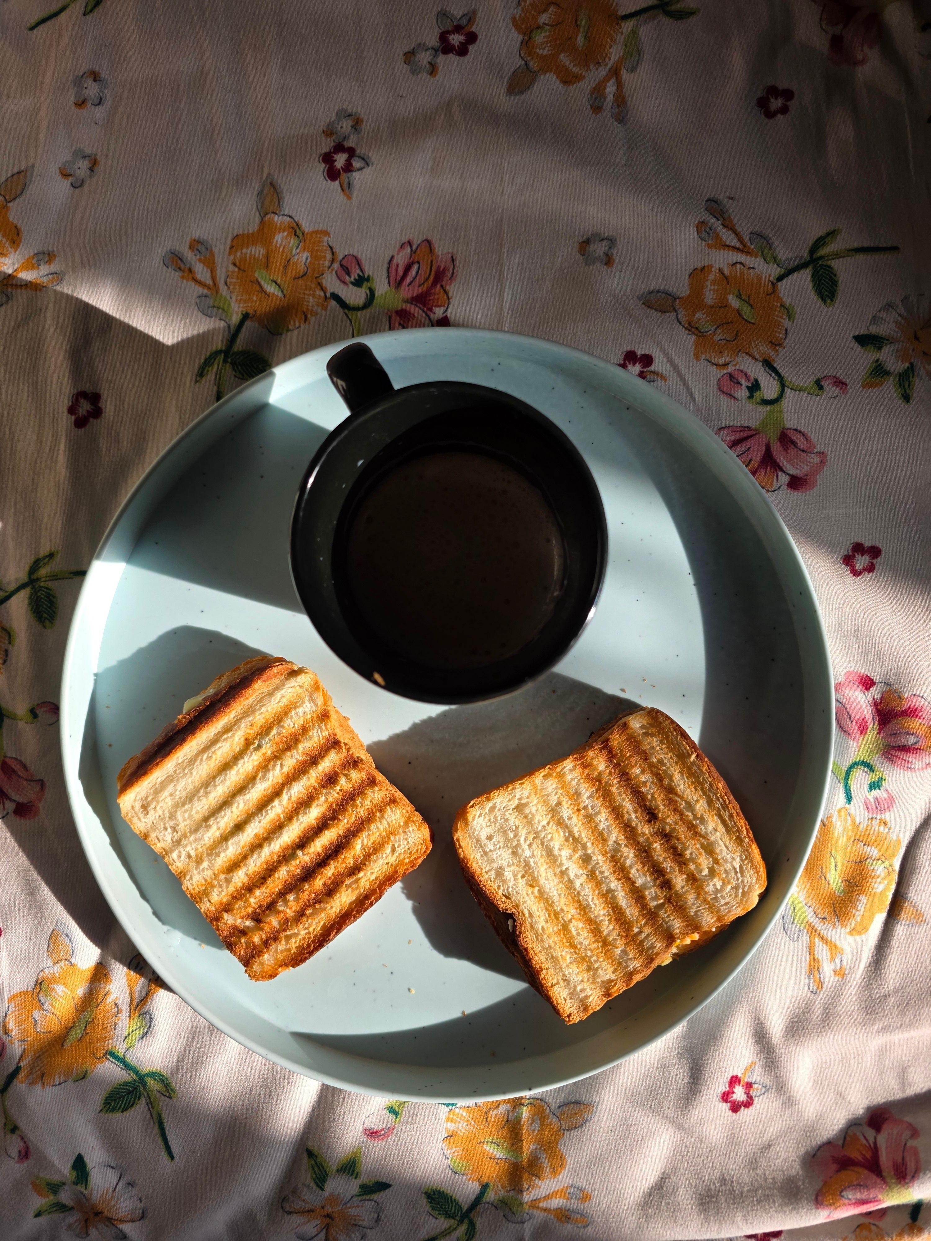 View of toasted sandwiches and coffee on a floral tablecloth, casting shadows.