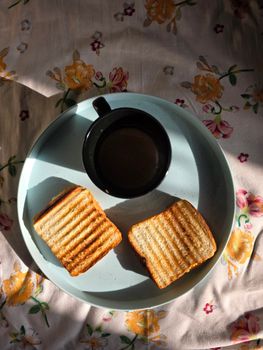 View of toasted sandwiches and coffee on a floral tablecloth, casting shadows.
