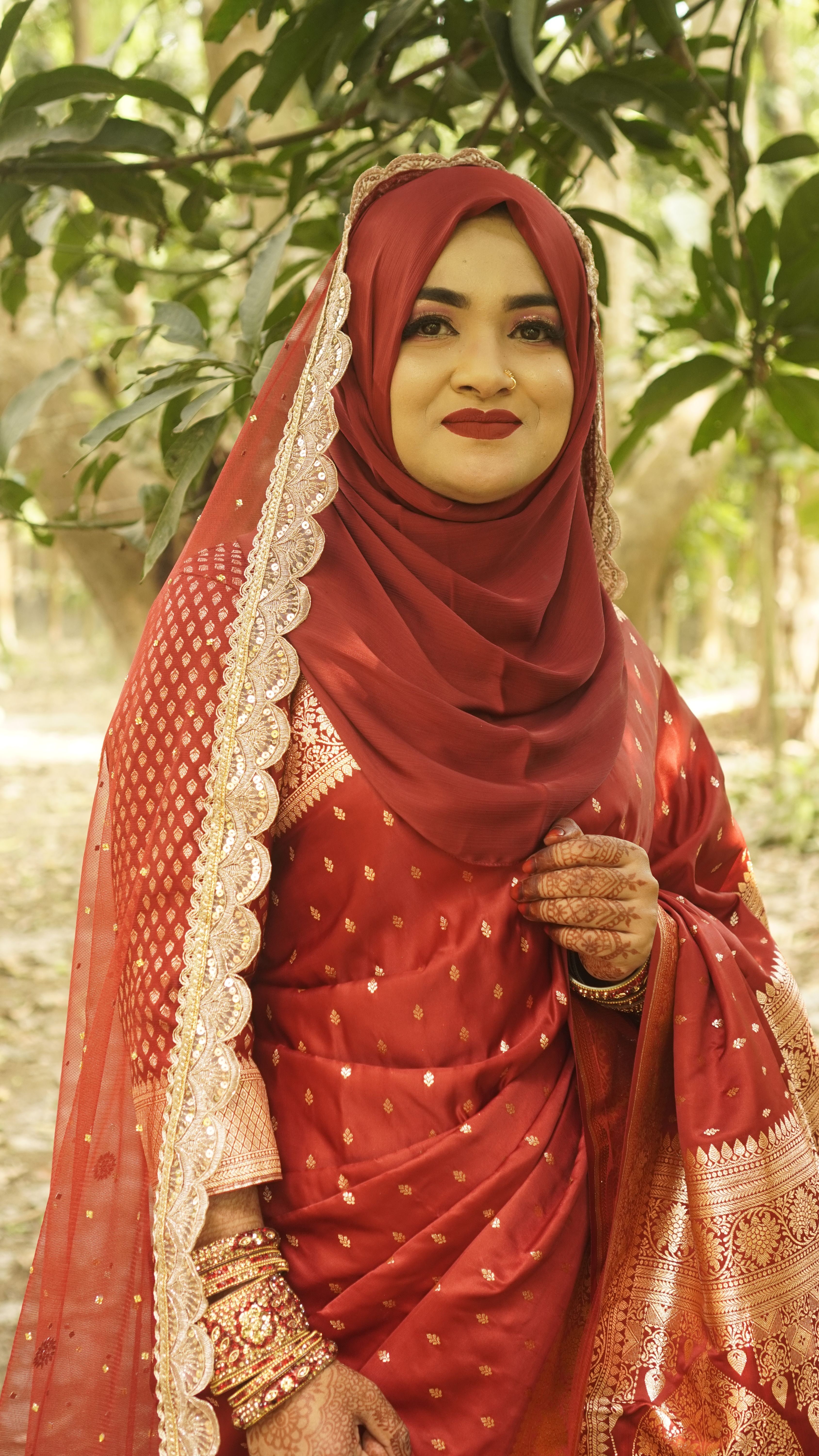 A woman in red traditional attire poses gracefully in a natural outdoor setting.