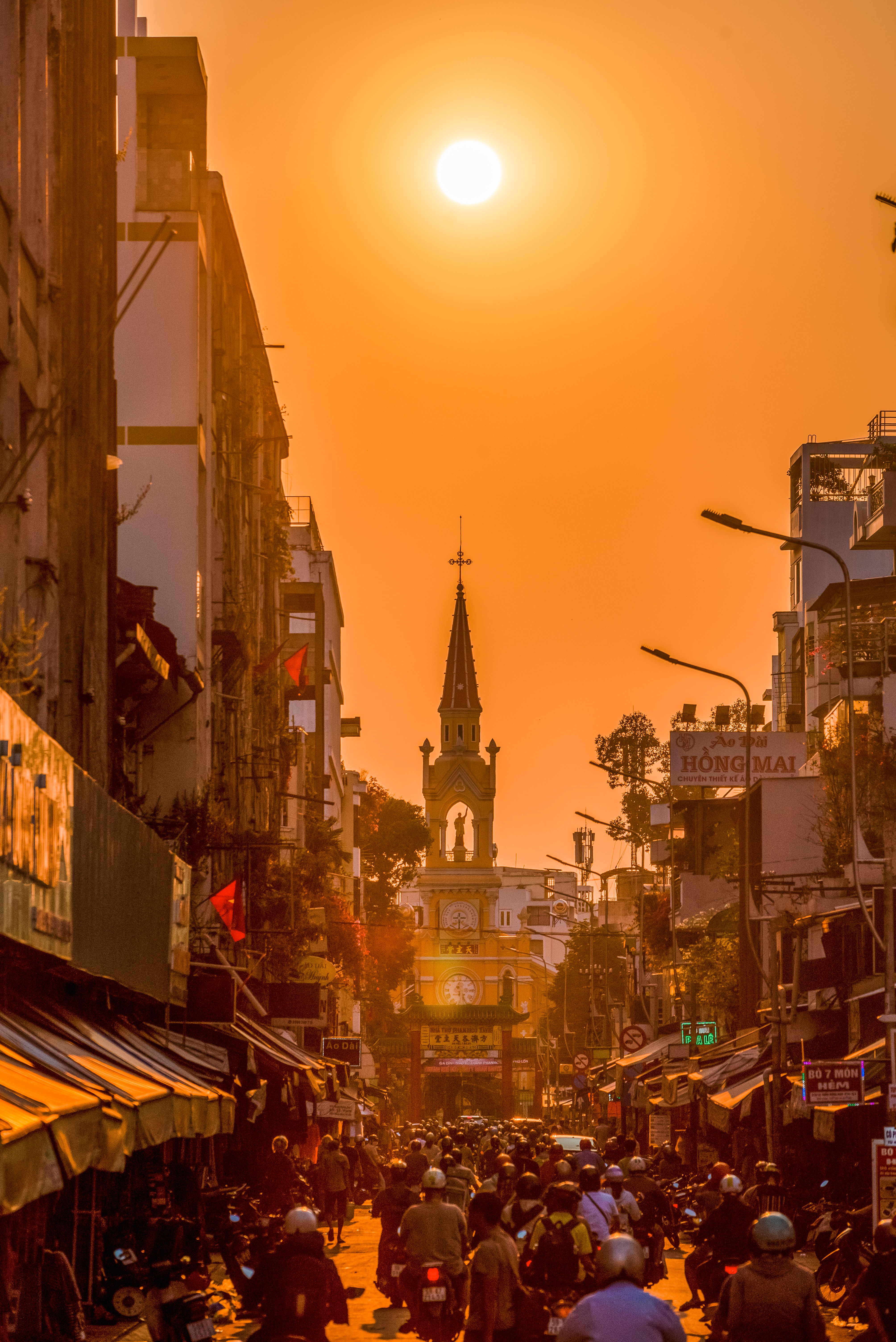 A vibrant sunset casts an orange glow over a busy street in Saigon, Vietnam, with prominent church architecture.