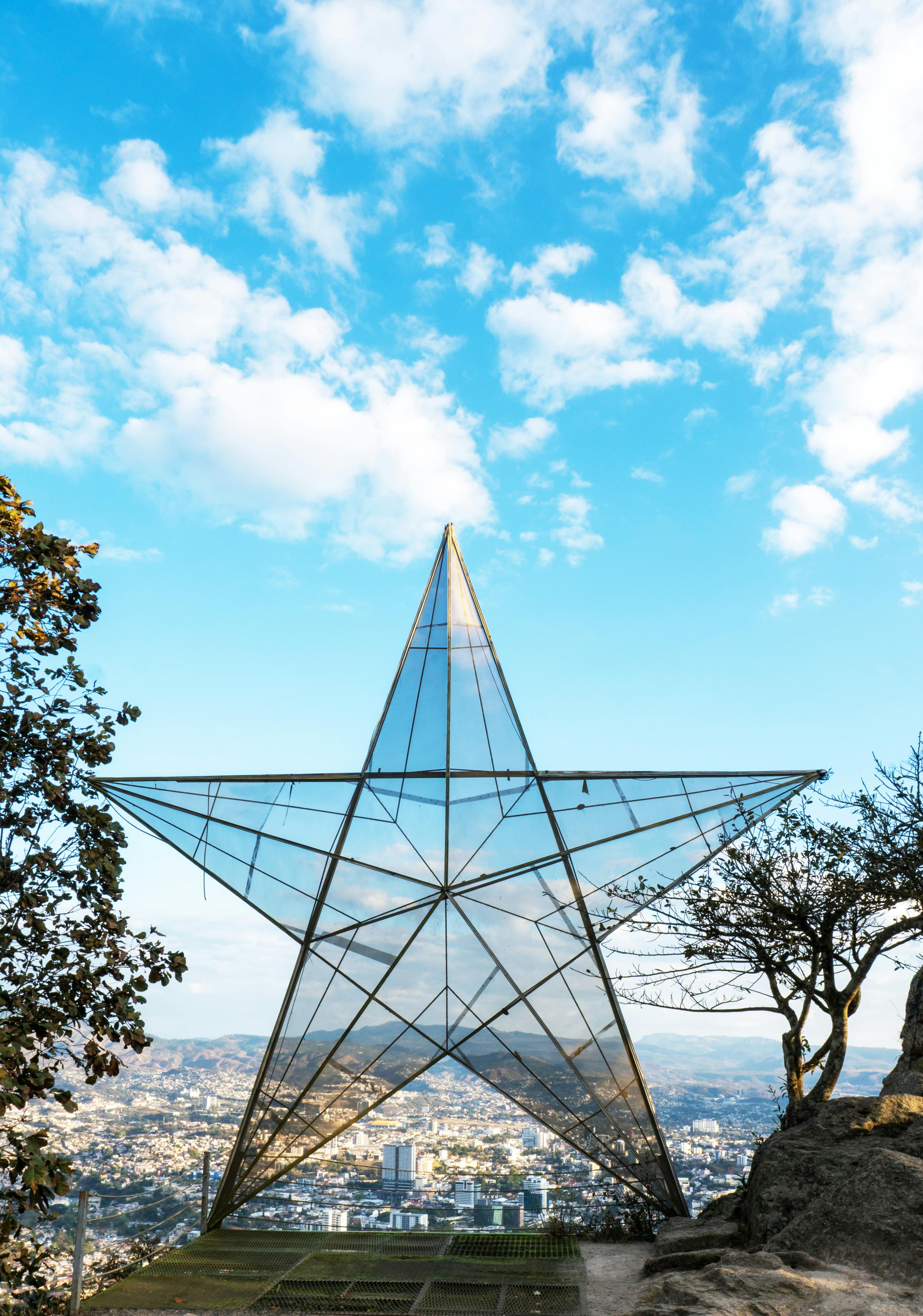 A glass star sculpture on a hill with a cityscape below under a bright blue sky.