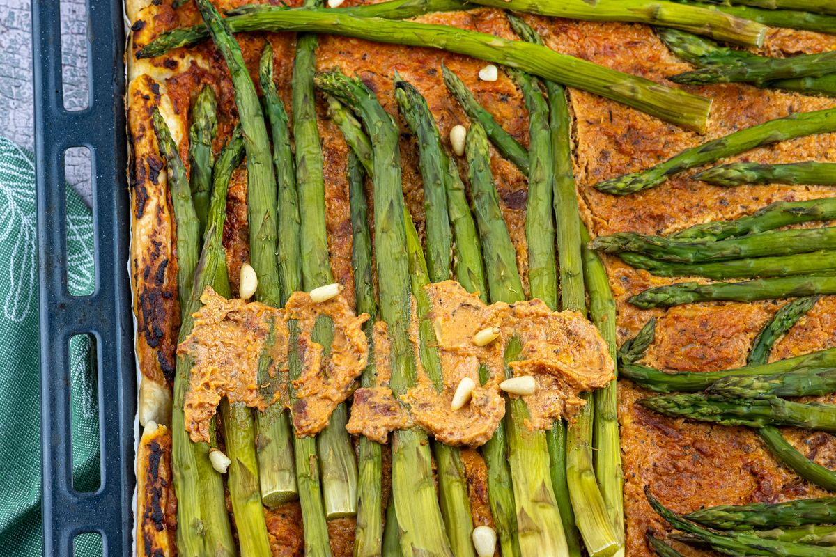 Sheet tart of green asparagus spears over red pepper spread with pine nuts on baking tray