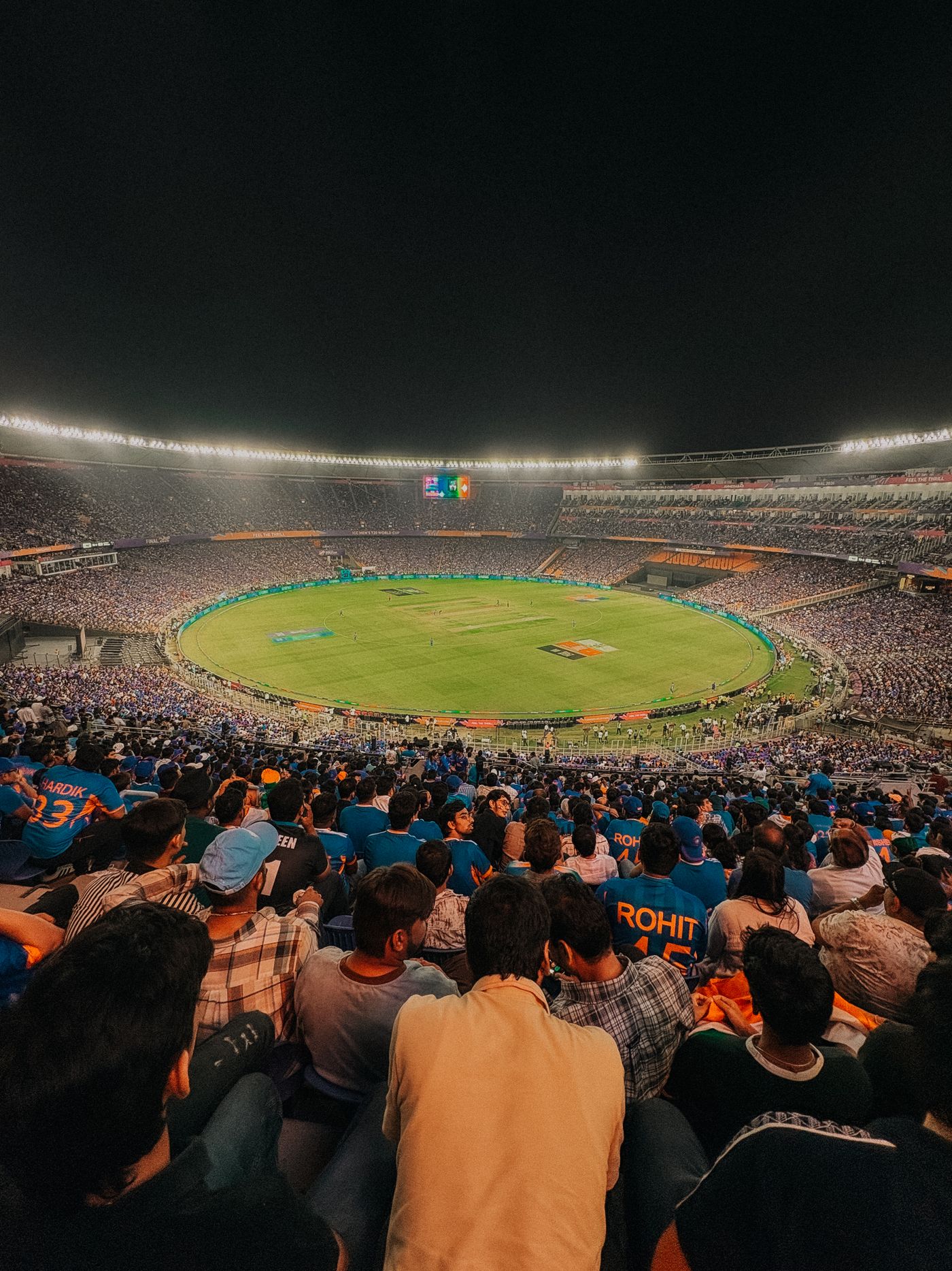 Packed cricket stadium at night under floodlights, fans in blue jerseys watching a match on the bright green outfield