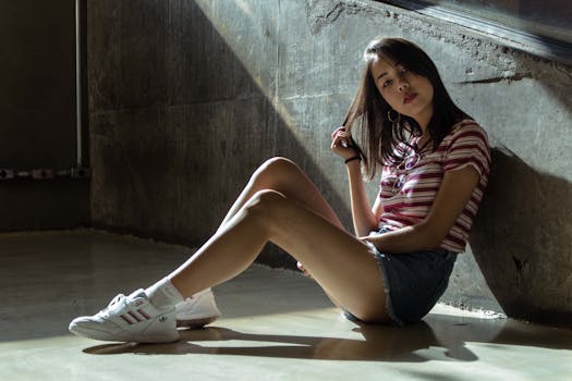 A young woman in casual wear poses elegantly indoors against a concrete wall.