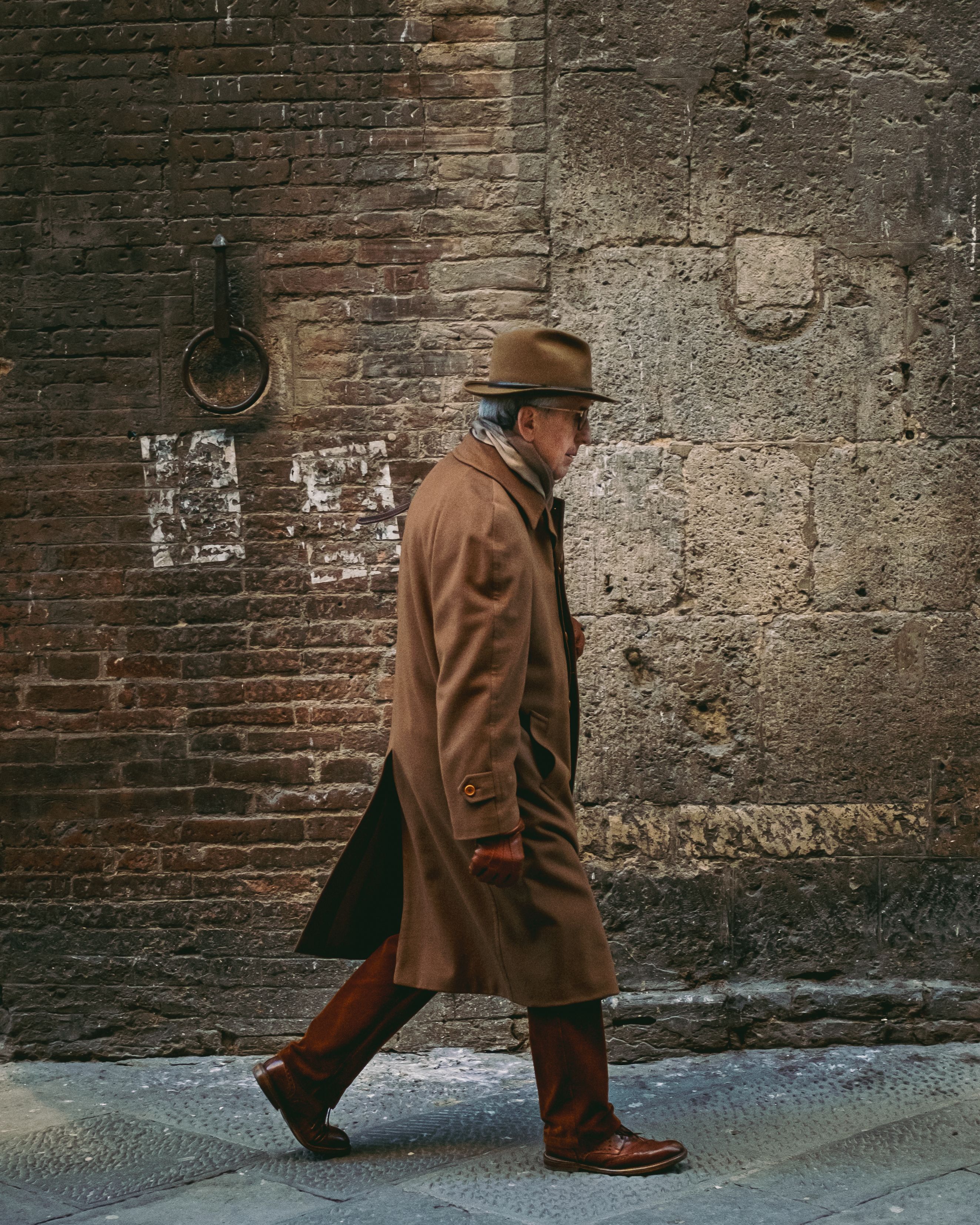 Elderly man in brown coat and hat walking along a historic brick street alley.