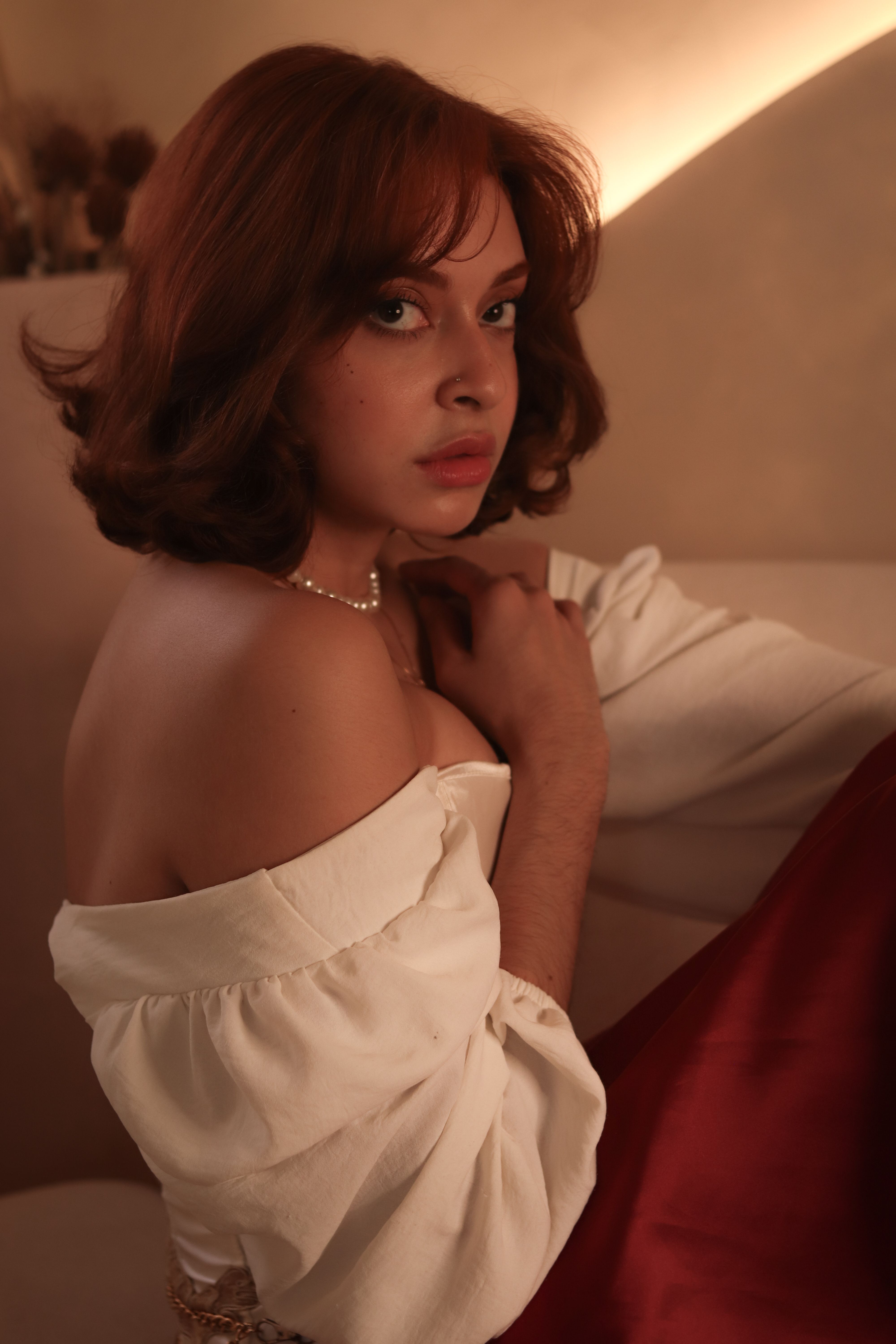 A woman in an off-shoulder top and red skirt poses gracefully, captured in warm indoor lighting.