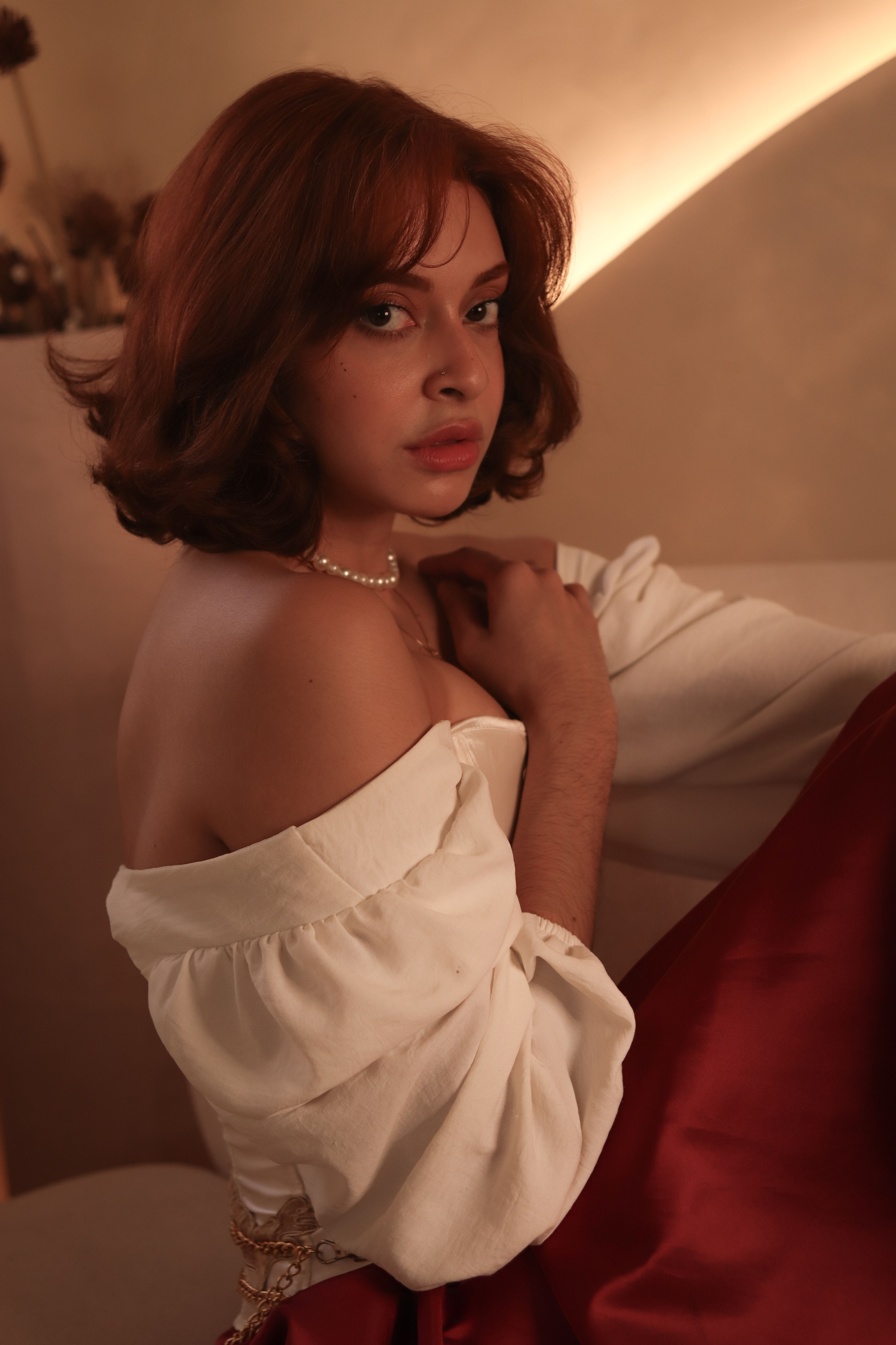 A woman in elegant attire poses indoors with a warm, moody ambiance highlighting her features.