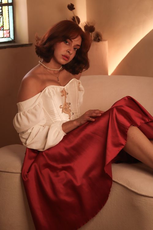 Free Woman with red hair in vintage off-shoulder dress sitting indoors on a couch with warm lighting. Stock Photo