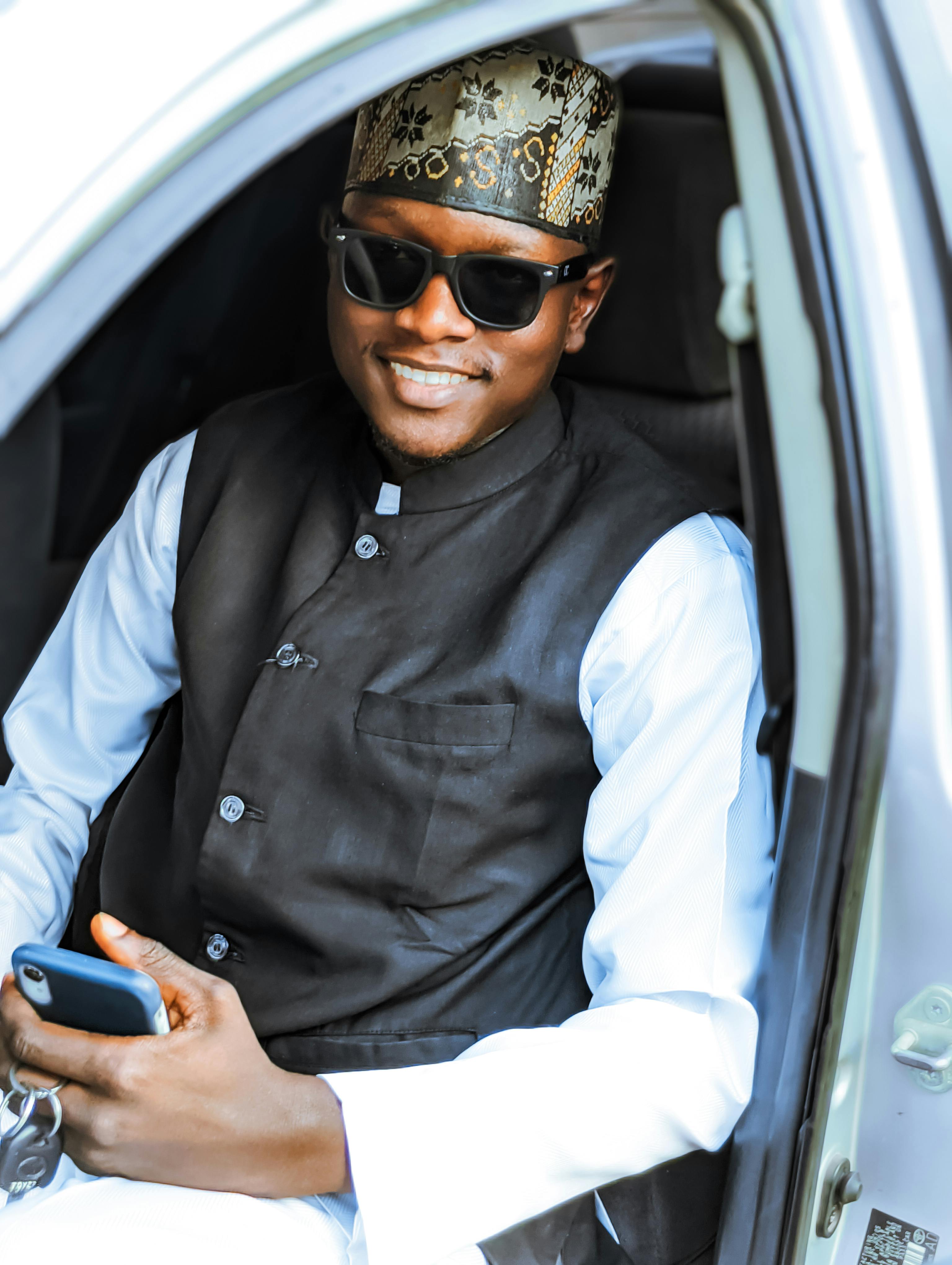 Well-dressed African man wearing sunglasses, holding a phone and smiling inside a car.