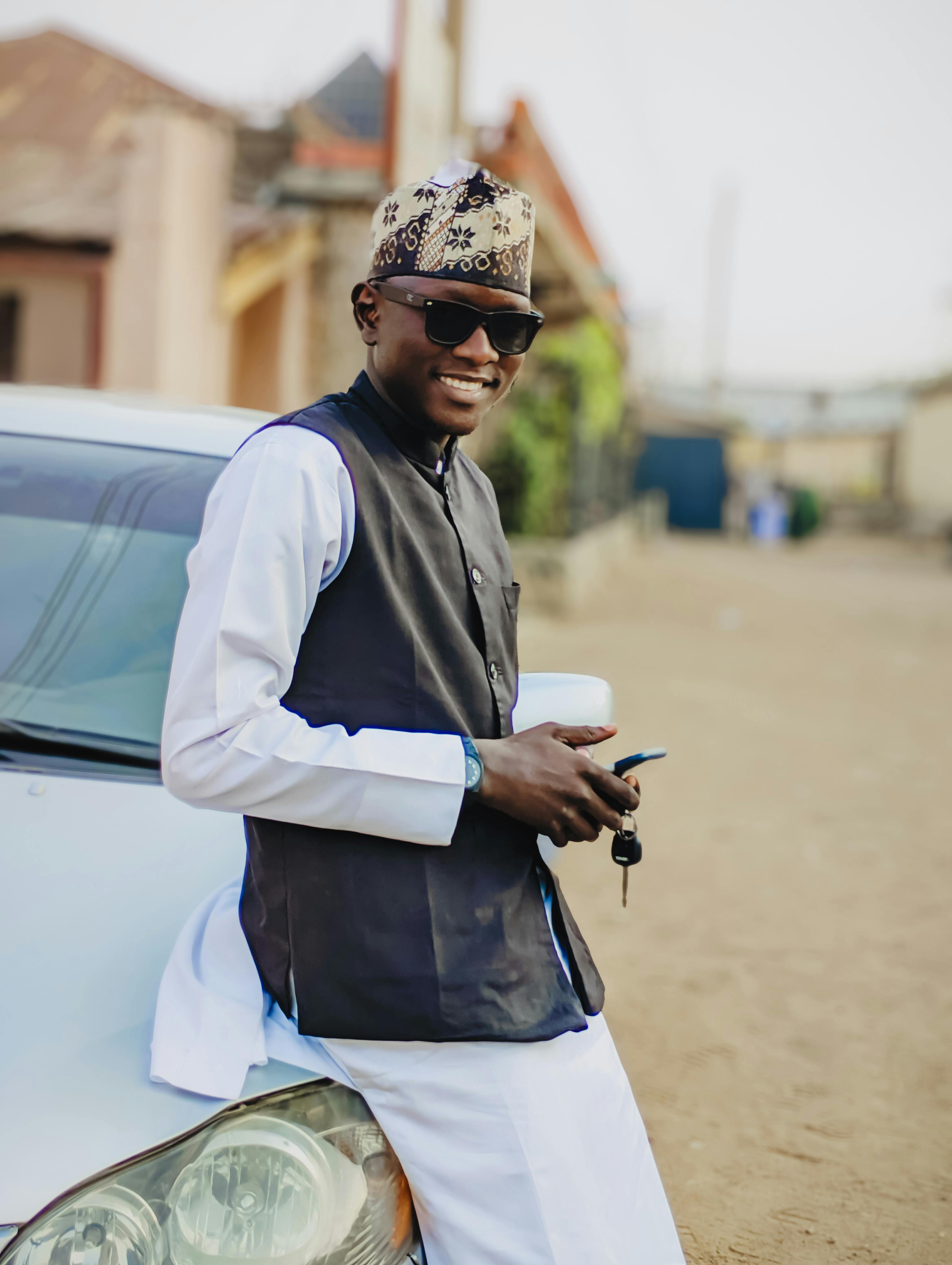 Smiling man in traditional attire leaning on a car outdoors, holding a phone and car keys.