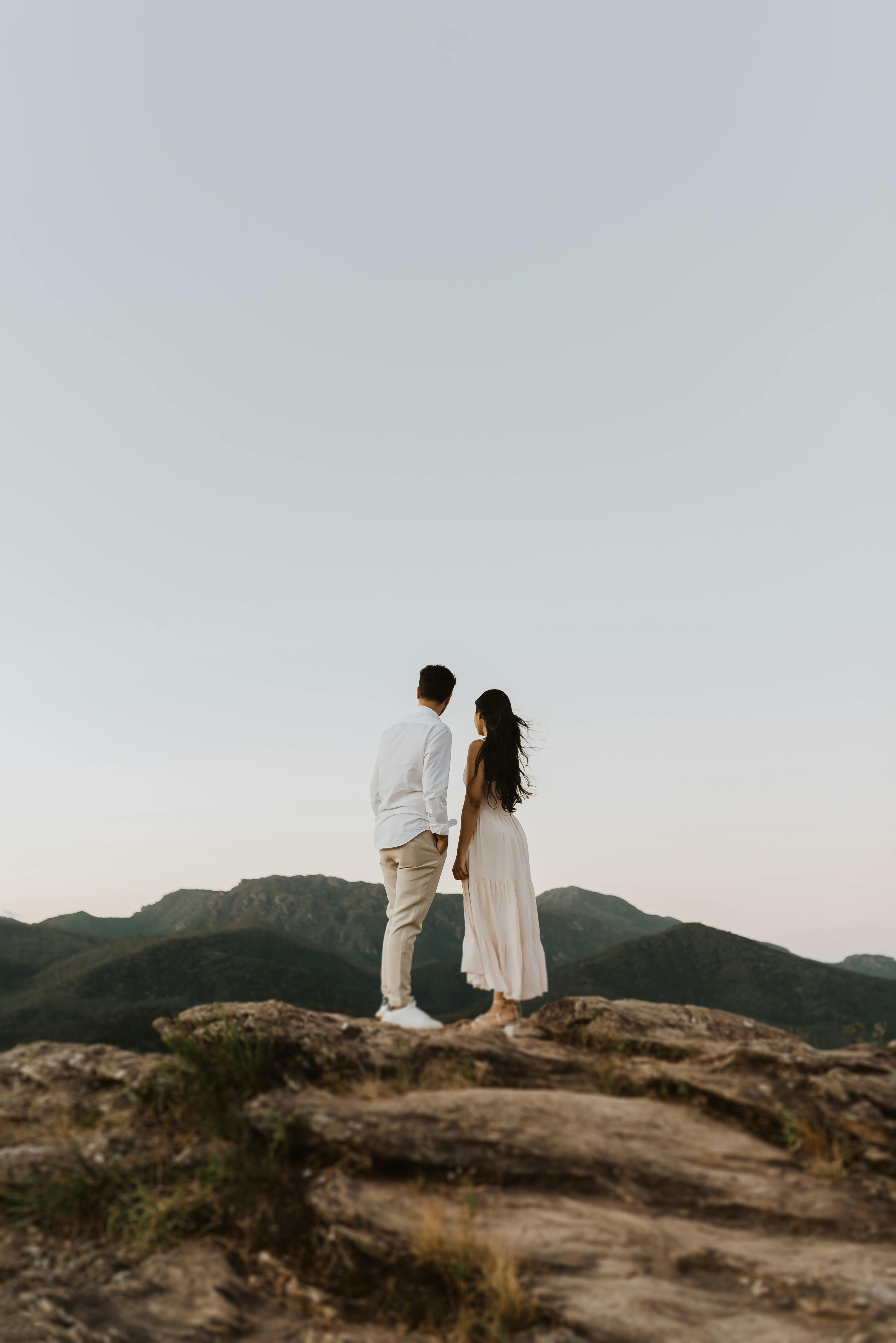 A couple stands on a rocky hilltop gazing at distant mountains under a serene sky.