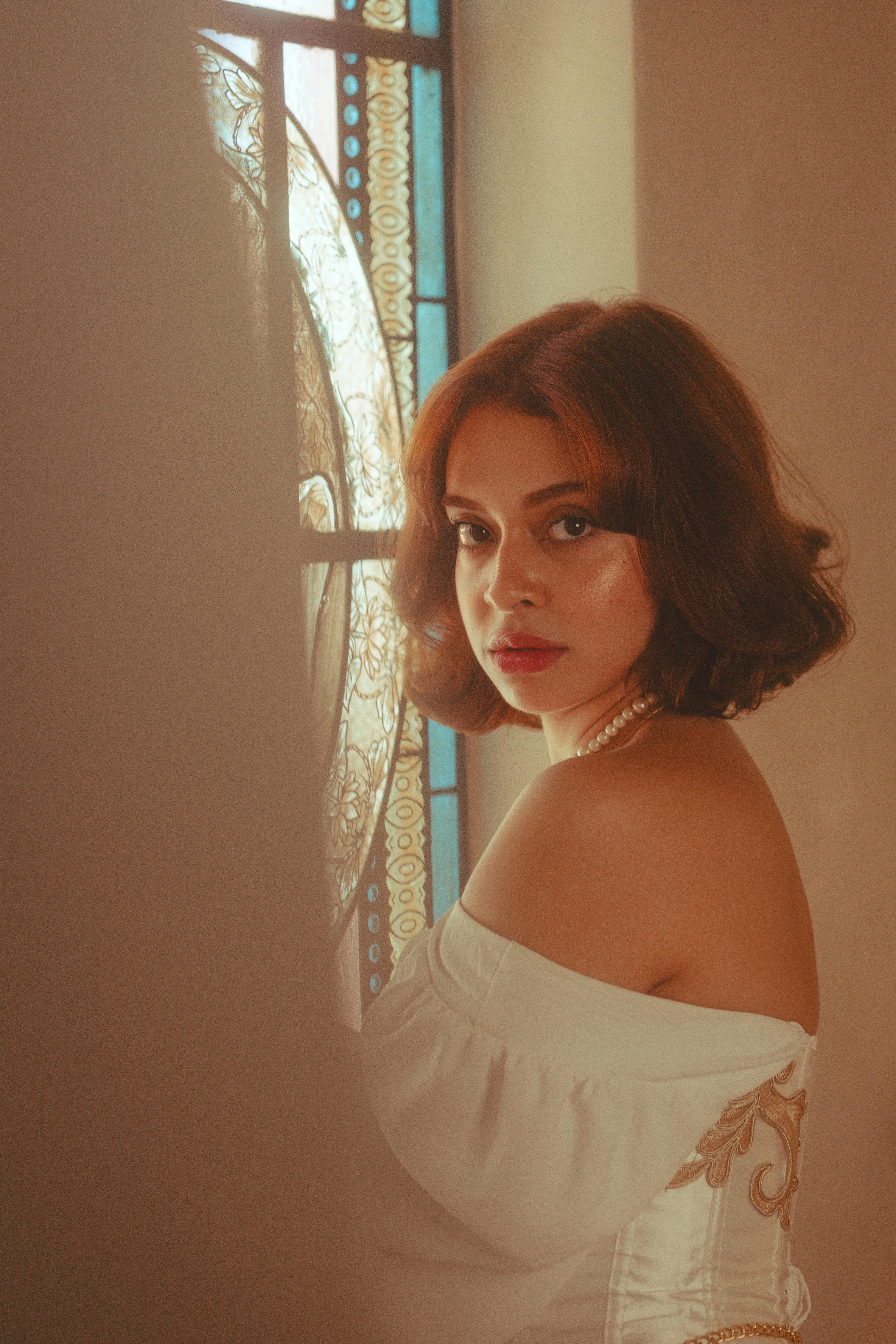 A stylish woman stands gracefully by a detailed stained glass window indoors.