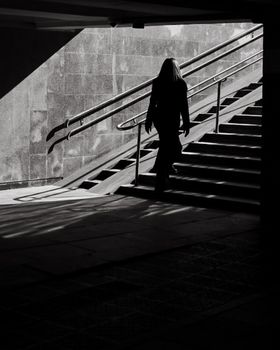 A woman in silhouette walks up stairs in a shadowy urban setting, black and white.