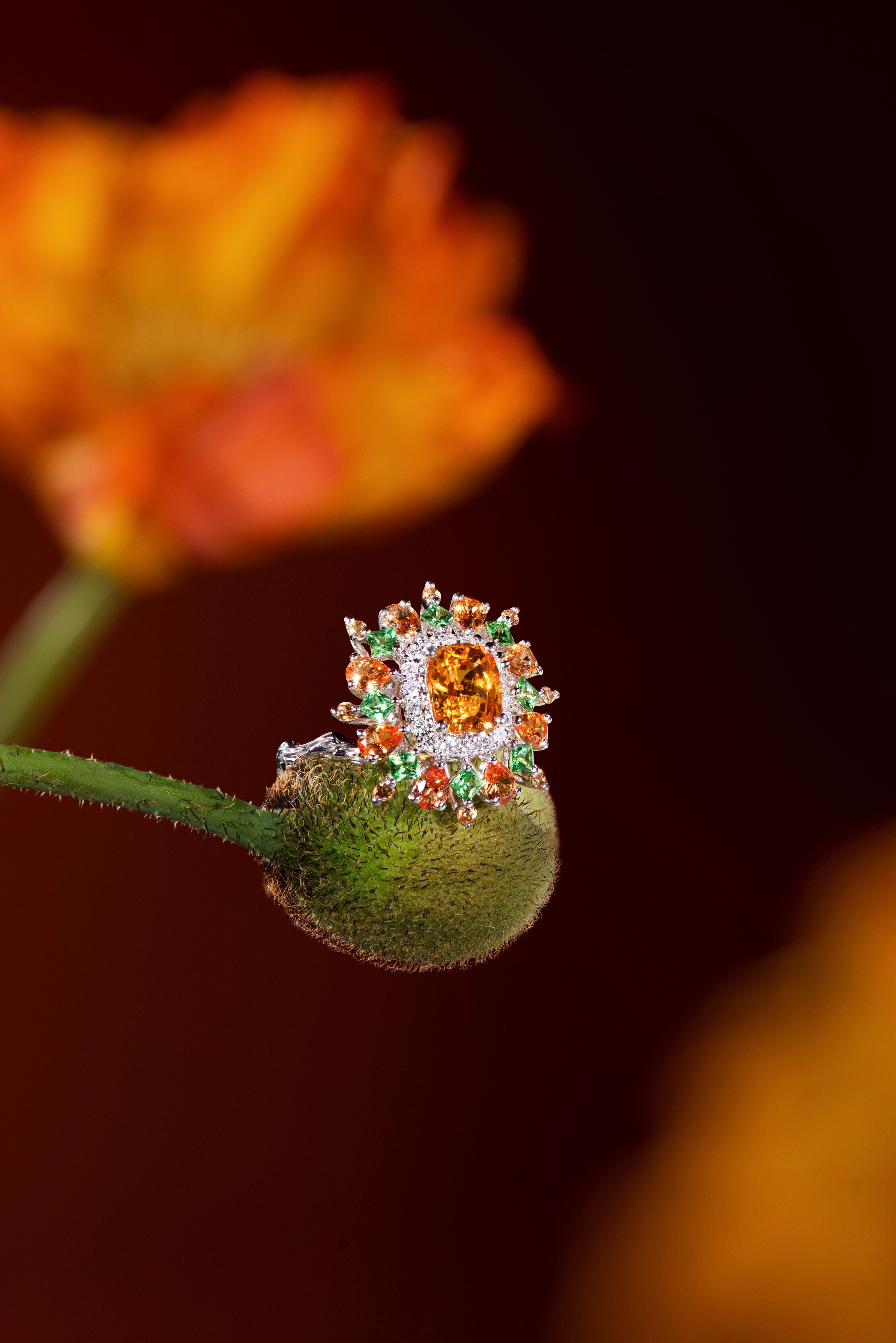 Intricate orange gem-studded ring gracefully perched on a green bud, evoking a floral essence against a warm, blurred background.