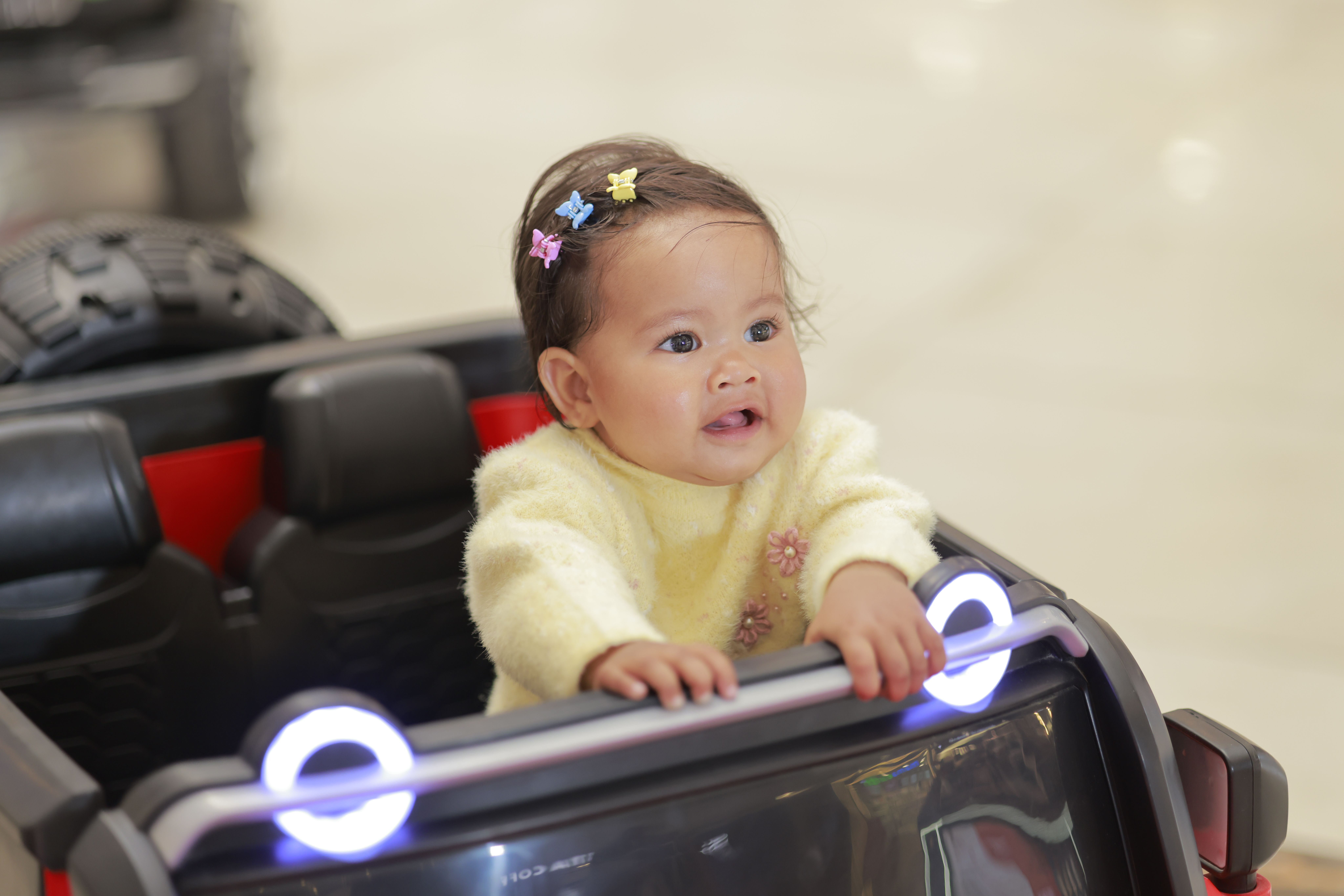 Cute baby in a toy car indoors, wearing colorful clips and a yellow sweater.