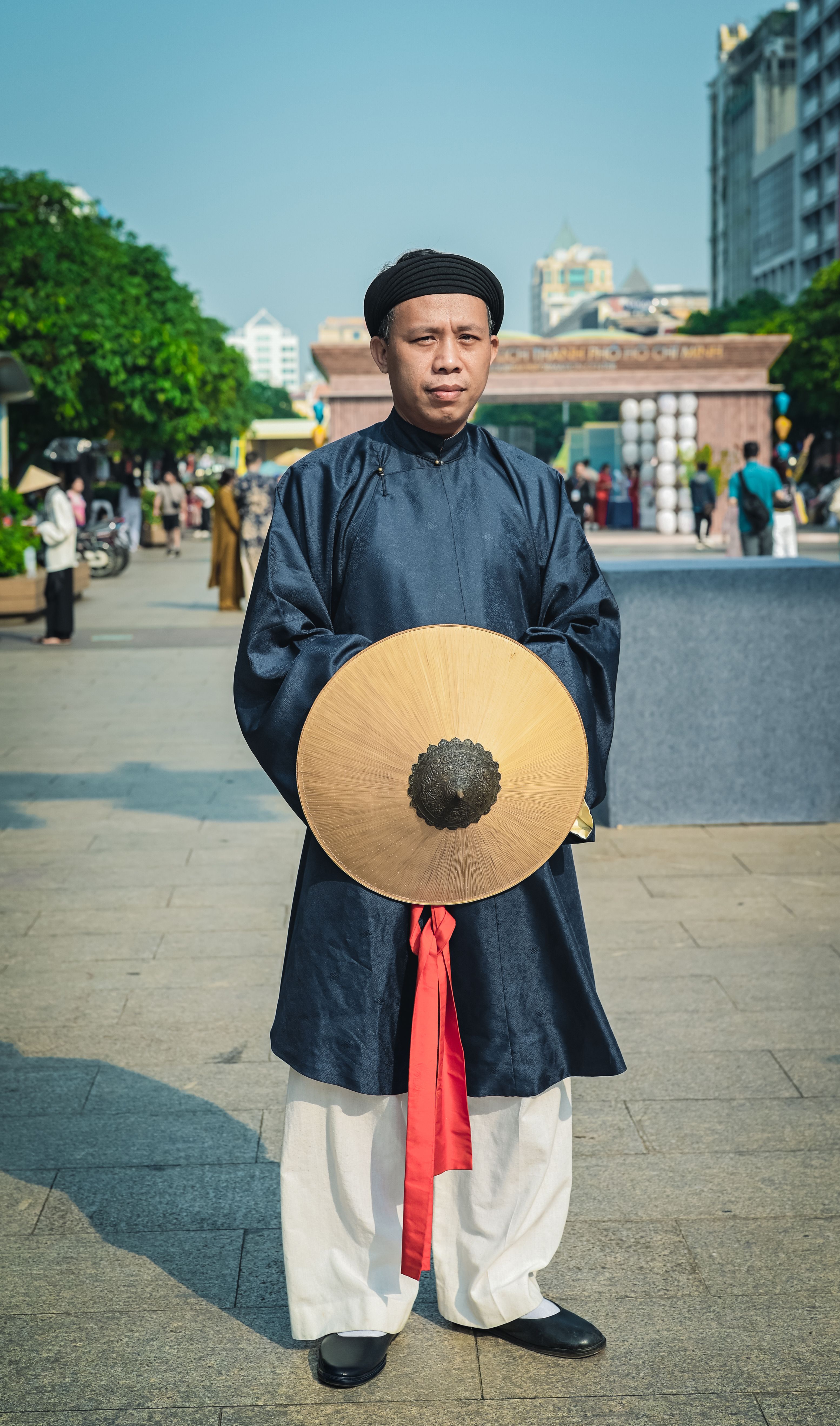 Man in traditional Asian attire holding a conical hat in a city setting.