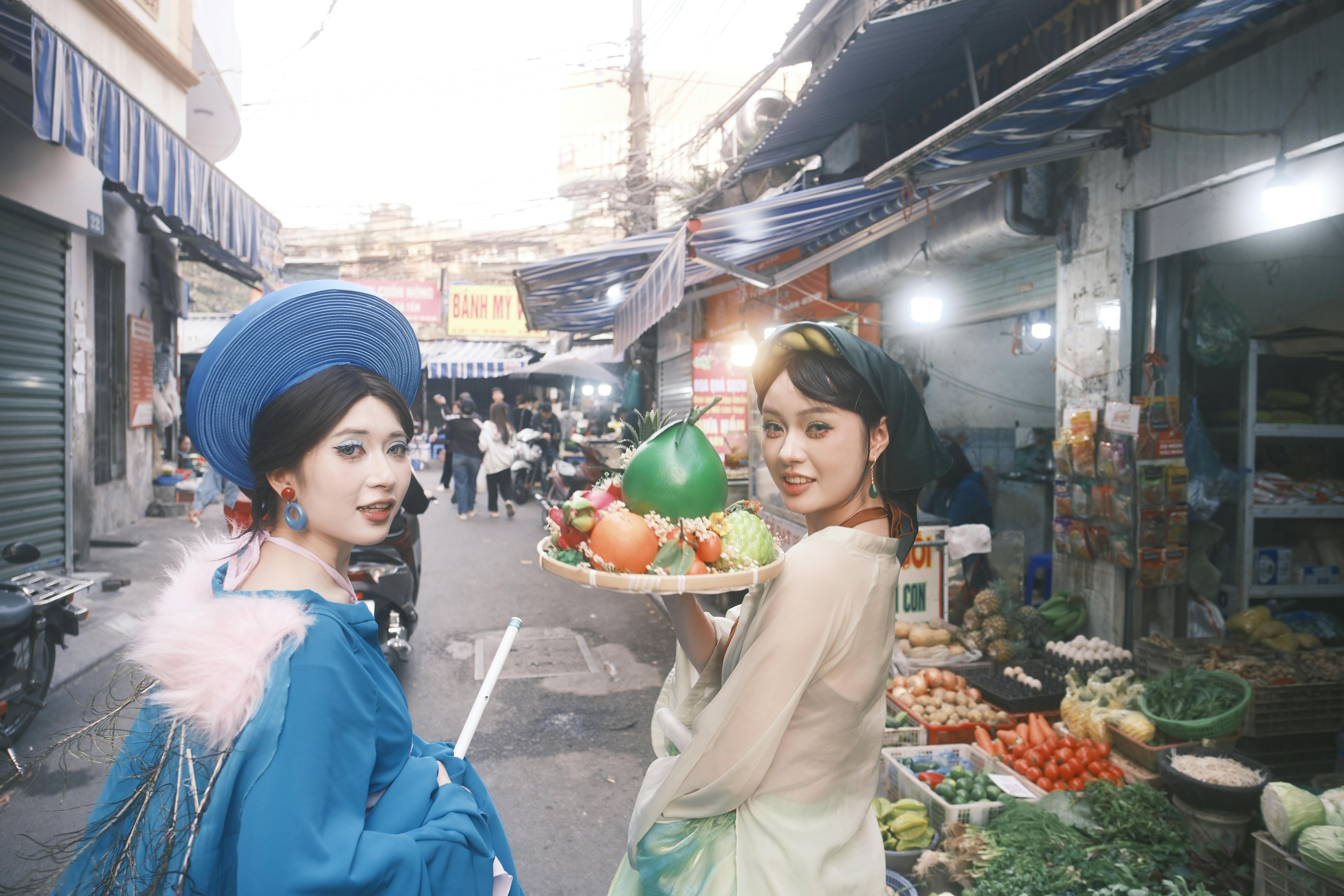 Two women in traditional attire pose with fruit in a vibrant street market setting.