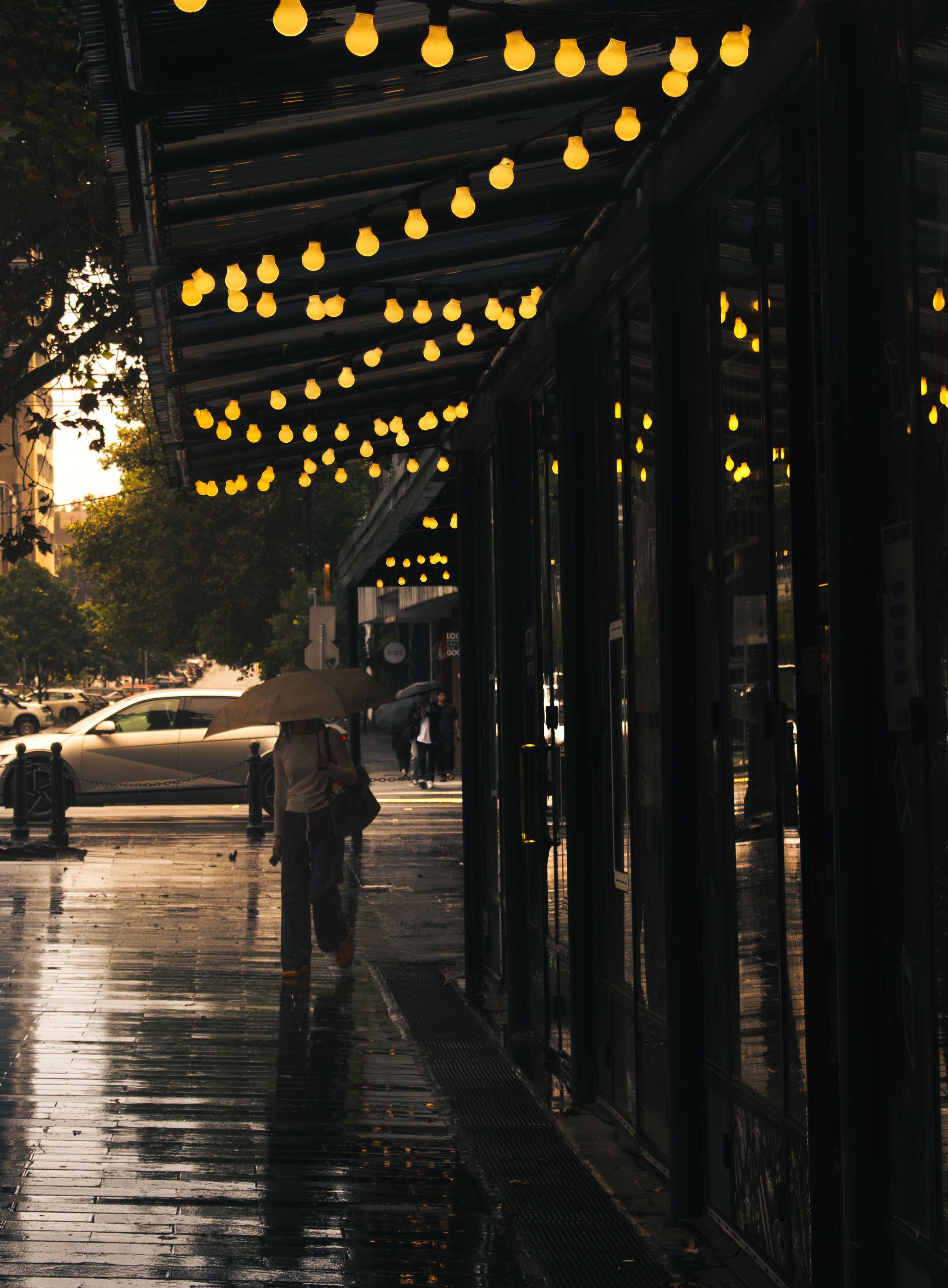 A person with an umbrella walks down a rainy city street in Melbourne, adorned with decorative lights.