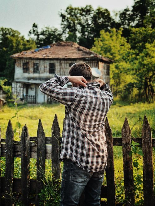 Free A man wearing plaid stands near a rustic, abandoned house surrounded by greenery. Stock Photo