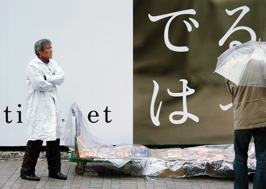 東京の街角で、雨の中、露天商と傘を差した通行人。対照的な日本の都市風景が描かれている。