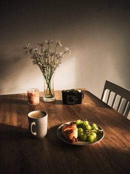 Warm morning light over a cozy breakfast with croissant, grapes, coffee, and flowers.